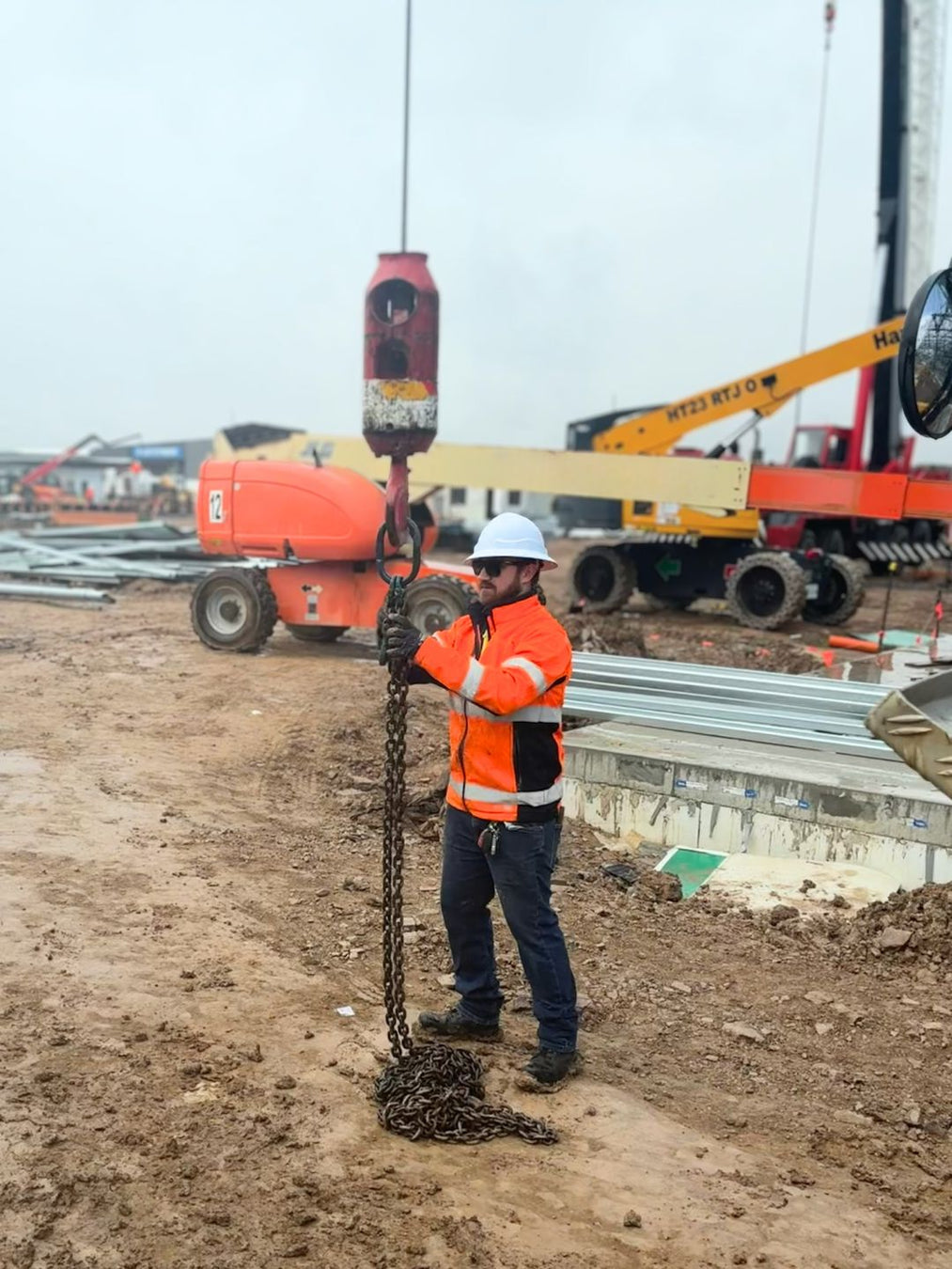 SLE technician in orange hi-vis and hard hat holding a chain sling on a construction site with an EWP and mobile crane